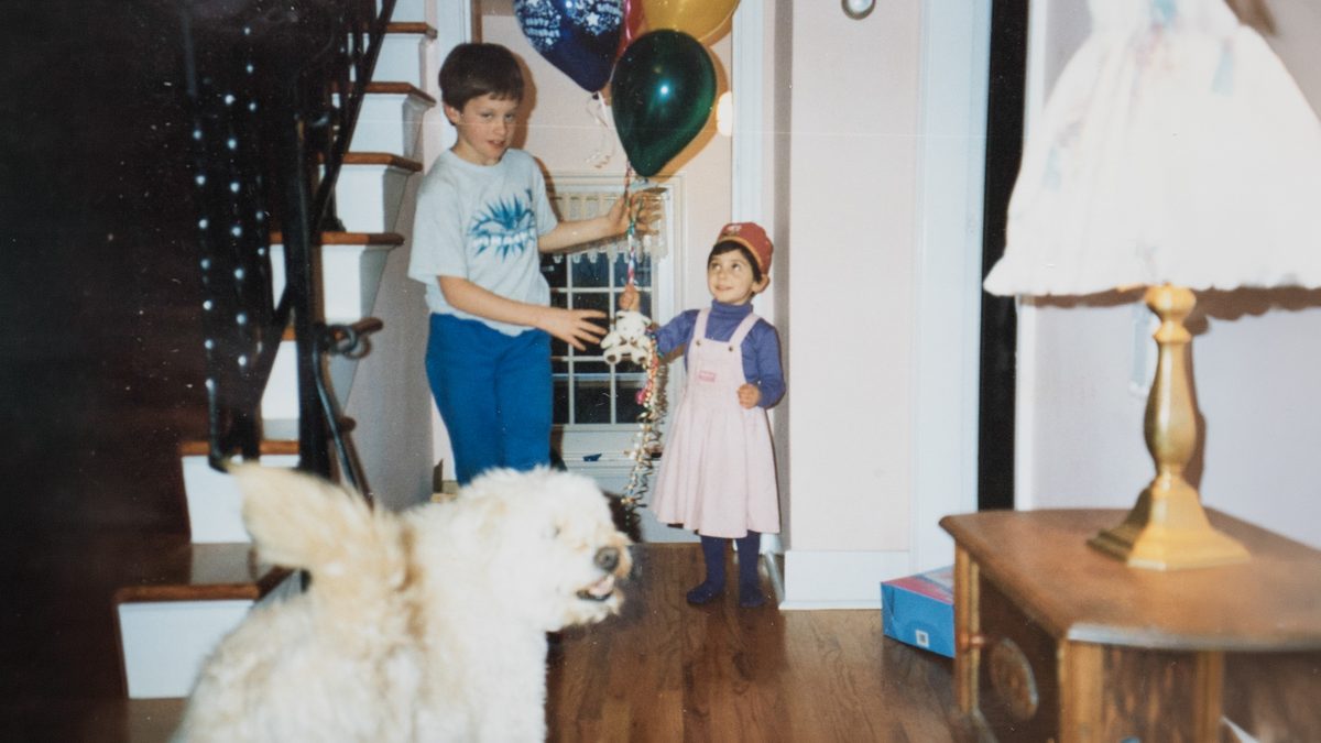 A boy holding balloons beside a small girl in a pink pinafore